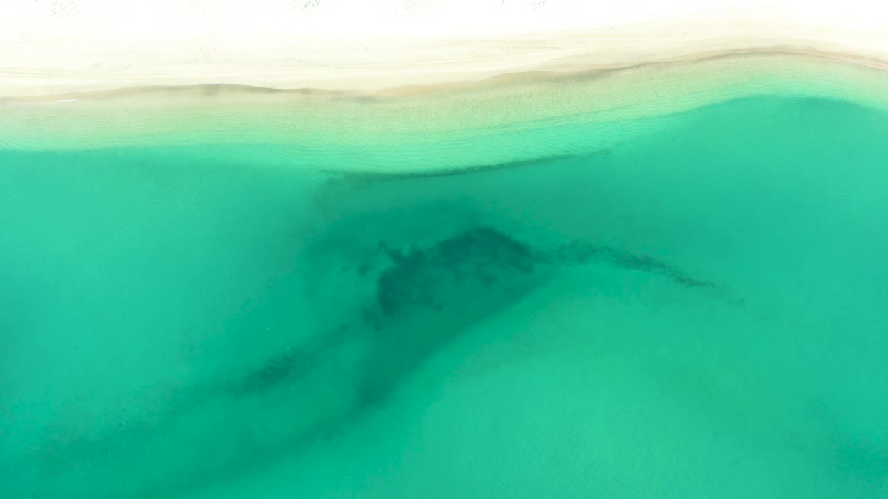 Top-down frame from La Ribera with the sandy beach running along the upper edge, green-blue water below, a soft dark patch under the surface, and no visible people.