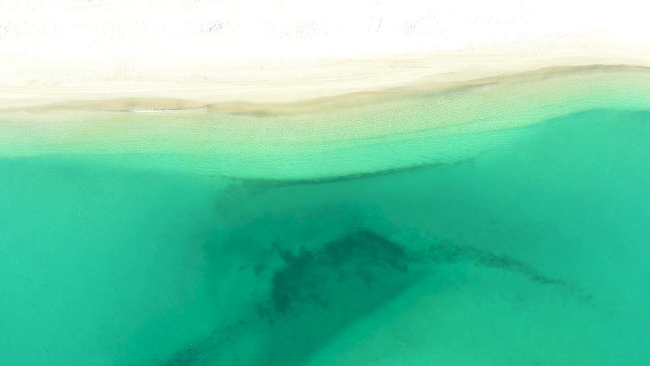 Aerial view of clear green water off La Ribera with a darker submerged patch below the pale shoreline and no visible people.