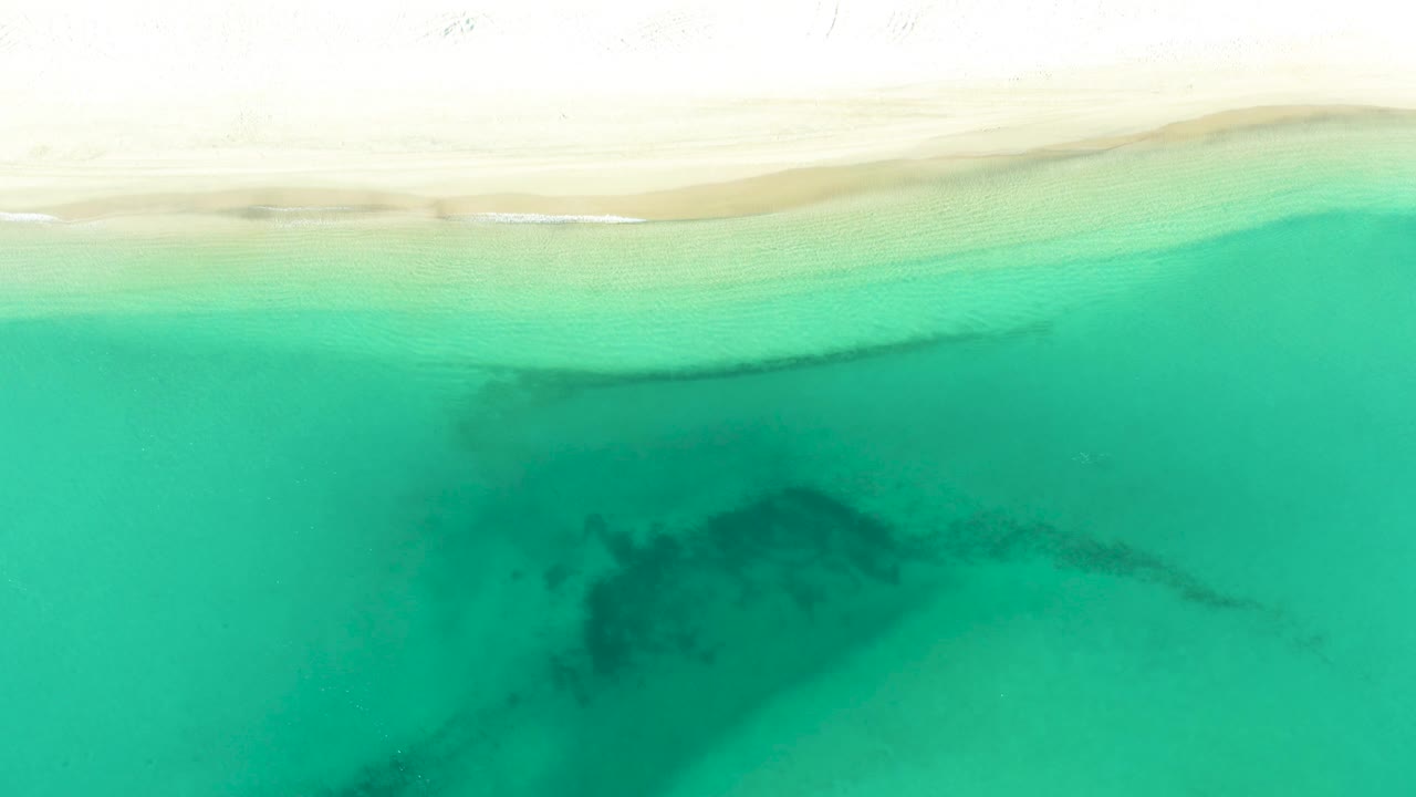 Aerial frame from La Ribera showing bright sand along the upper edge and clear green water with a darker area drifting through the lower frame, no visible people.