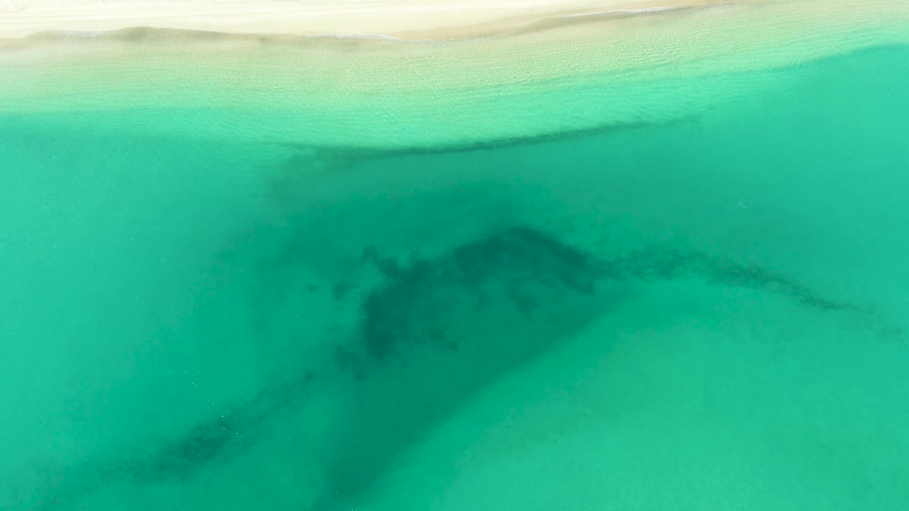Top-down view of La Ribera coastal water where a dark submerged shape sits below the turquoise surface and the pale beach remains visible along the upper edge, no visible people.
