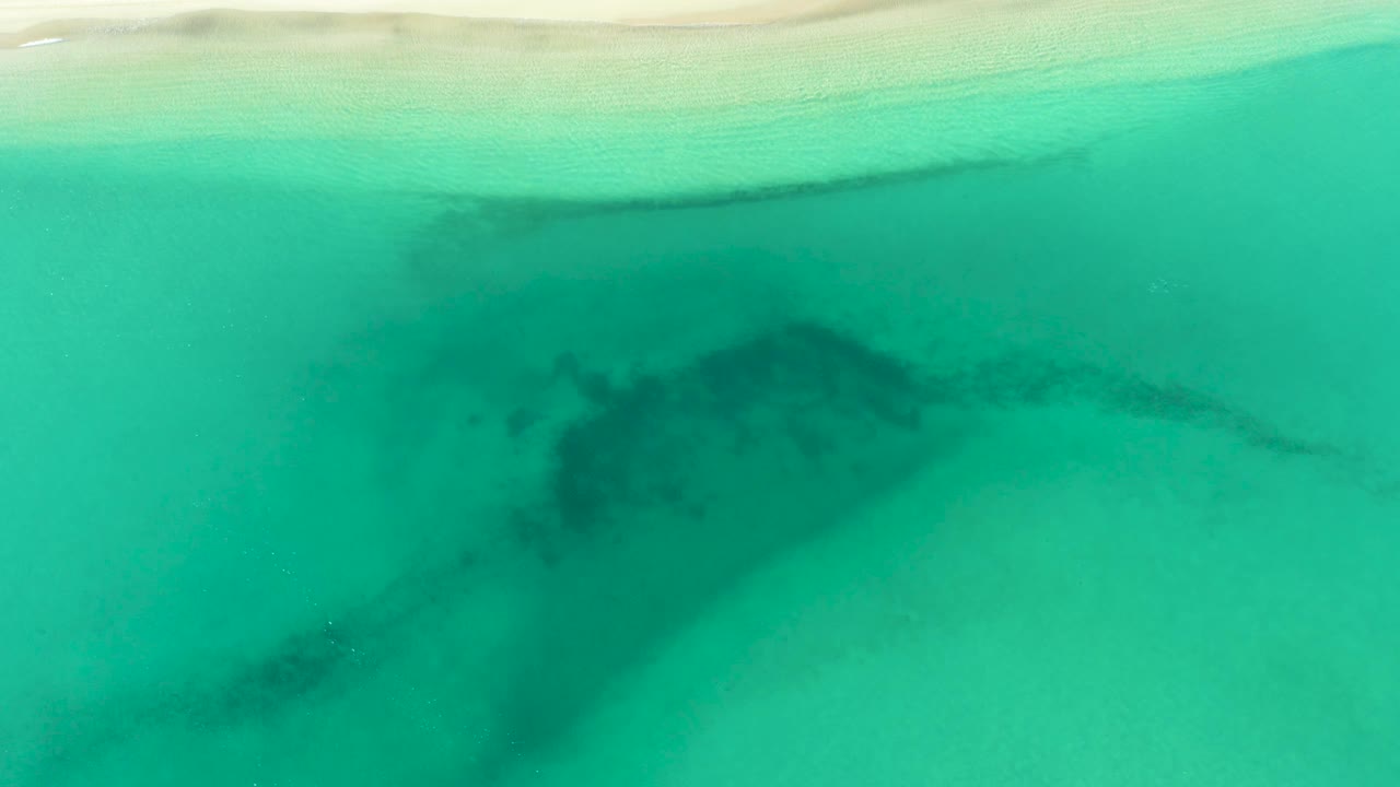 Aerial view over La Ribera shallows with a wider turquoise field, pale sand at the top, and a dark underwater patch crossing the center-left area, no visible people.