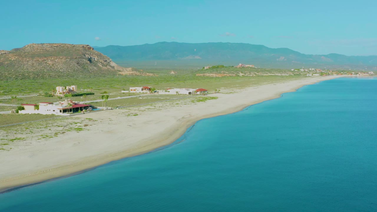Opening aerial frame over La Ribera, Mexico, with turquoise water at right, pale beach curving left, low beachfront buildings, rocky hill, distant mountains, and no visible people.