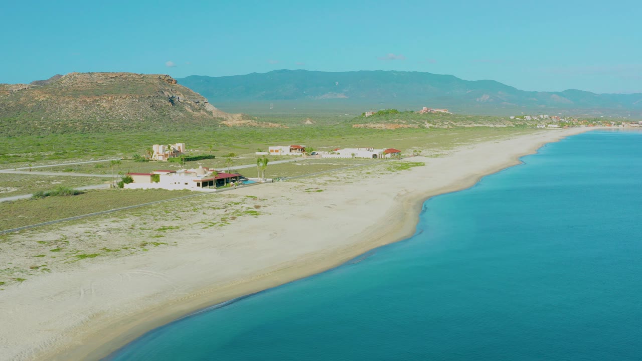 Aerial frame over La Ribera, Baja California Sur showing pale sand, turquoise shoreline, scattered coastal homes, dirt roads, scrubland, low hills, distant mountains, and no visible people.