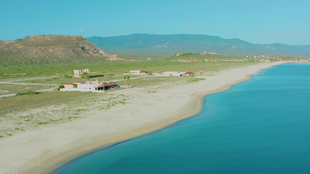 Wide aerial shoreline with houses, sand, blue water, and hills.