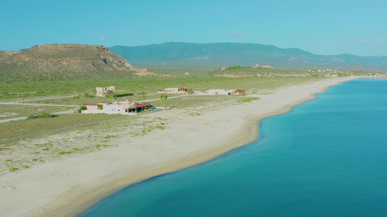 Aerial frame over La Ribera, Baja California Sur with a rocky hill on the left, small homes near the sand, a pale beach edge, turquoise water to the right, and no visible people.