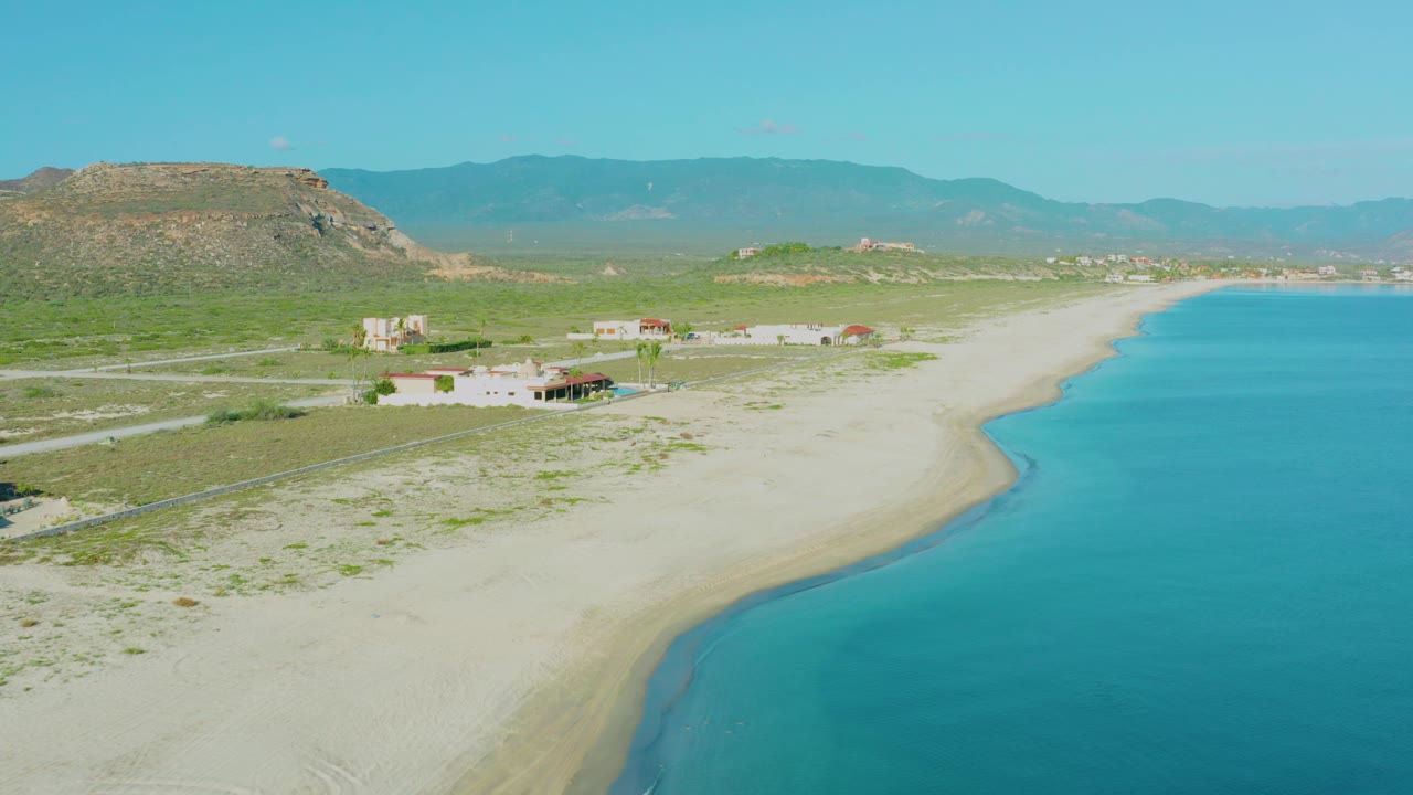 Aerial frame in La Ribera showing the shoreline running diagonally past turquoise water, small homes set back from the beach, inland roads, a flat-topped hill, and no visible people.