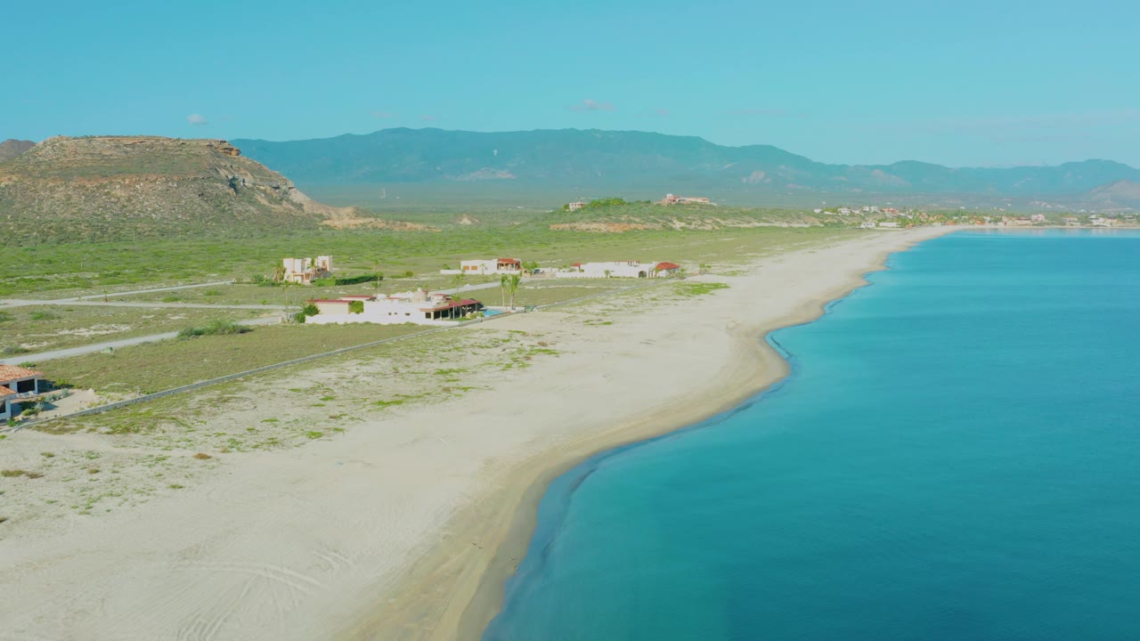 Coastal aerial with beach, sea, homes, access roads, hills, and mountains.