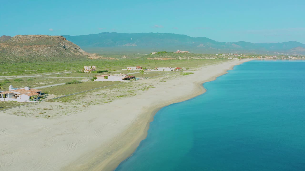 At 0:01, the La Ribera aerial view keeps the curving beach and clear blue water in frame, with beach houses and desert hills behind and no visible people.