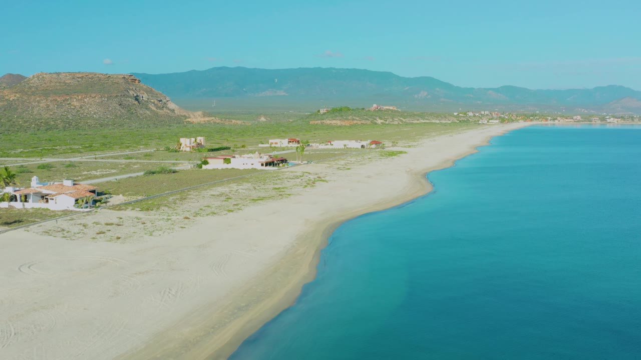 At 0:02, the frame shows the La Ribera shoreline from above with more open turquoise water on the right, pale sand below, low homes inland, and no visible people.