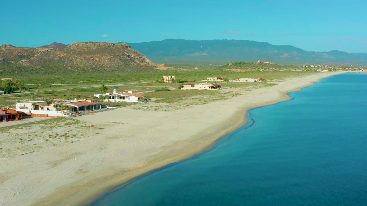 Aerial view in La Ribera, Baja California Sur, with pale sand and blue water beside low beach homes and mountains in the distance, no visible people.
