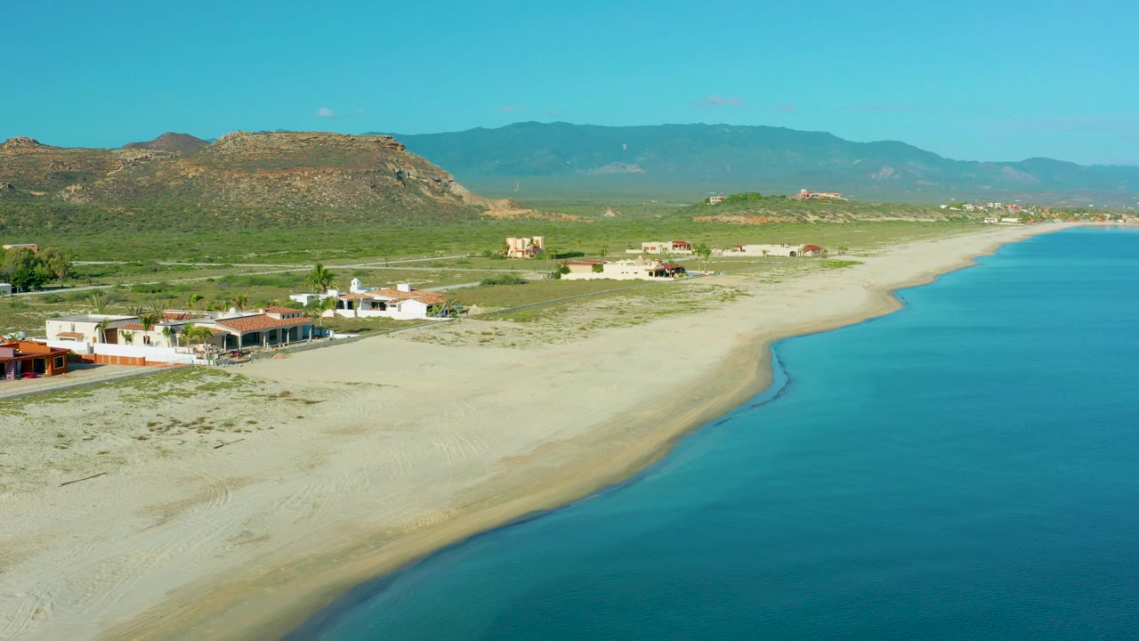 Aerial coastal view with sandy beach, nearby homes, blue sea, and hills.