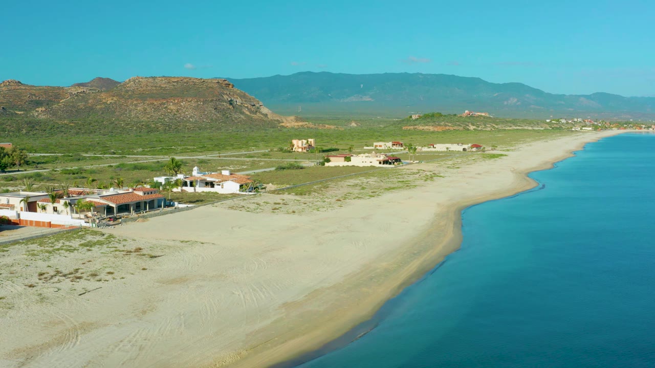 Wide aerial view in La Ribera with the curved shoreline on the right, soft sand across the center, coastal houses on the left, layered hills in back, and no visible people.