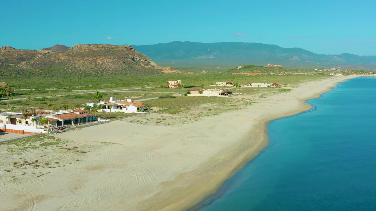 Drone-height view of La Ribera beach with turquoise water filling the right edge, broad pale sand in the middle, scattered homes to the left, hills behind, and no visible people.