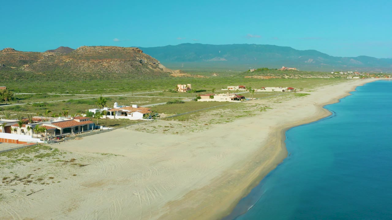 Elevated coastal view with houses near sand, blue water, and mountain backdrop.