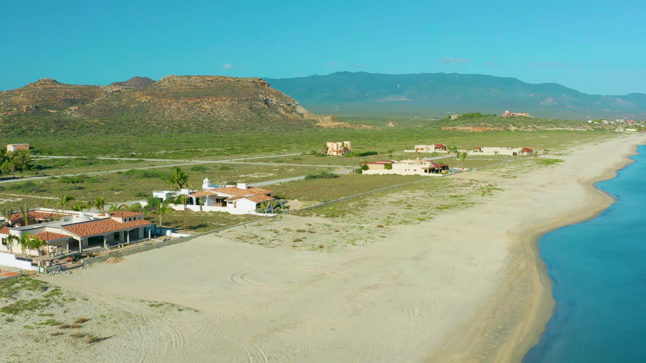 Wide aerial view of low buildings, sandy beach, blue water, and mountain horizon.