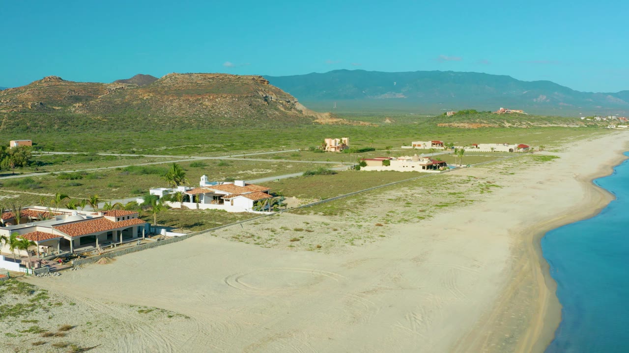 La Ribera coastline appears from above with terracotta roofs at the lower left, pale beach running into the distance, a strip of blue sea at right, scattered inland buildings, rocky hills, and no visible people.