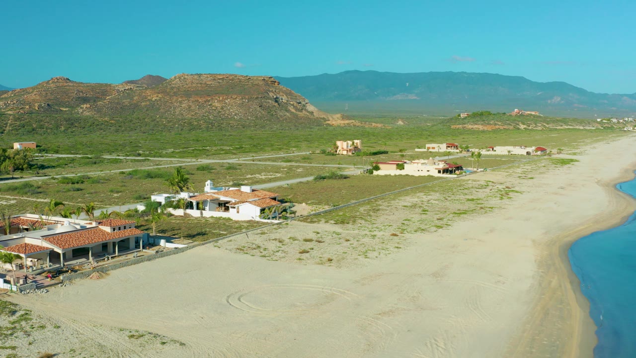 Drone view of beach homes at left, open sand, roads, scrubland, and blue water.