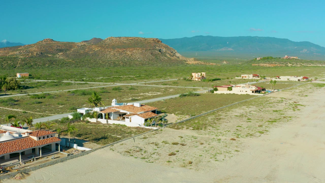 Aerial view in La Ribera, Baja California Sur, showing sandy open lots, red tile roof homes, roads, green scrub, a rocky mesa, distant mountains, and no visible people.
