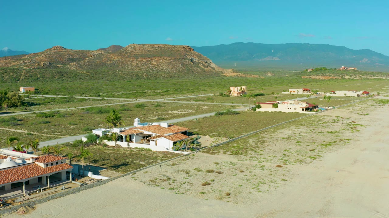 Aerial frame in La Ribera, Baja California Sur, with white houses and red roofs in the lower frame, pale sand lots, crossing roads, green scrub, mesa terrain, and no visible people.