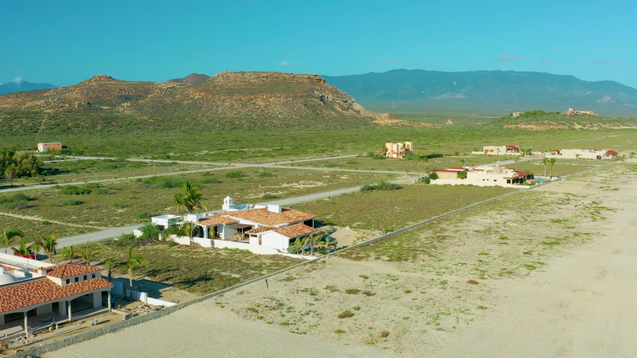 Aerial view in La Ribera, Baja California Sur, showing empty sandy plots beside low houses, a light road network through scrub, a rocky mesa, distant mountains, and no visible people.