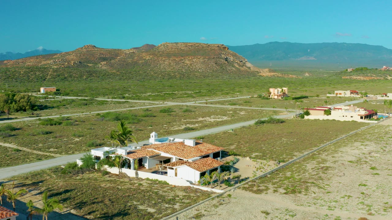 Opening aerial frame over La Ribera, Baja California Sur, with white homes and red tile roofs in the lower foreground, pale roads crossing green scrubland, distant hills, and no visible people.