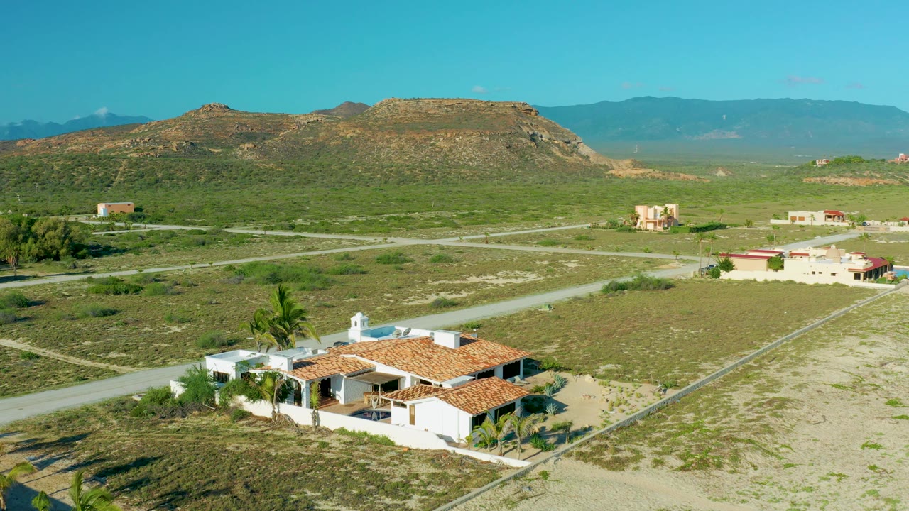 Third frame over La Ribera places the tile-roof home at the lower right edge, with a pale road running diagonally through green land toward low rocky hills and no visible people.