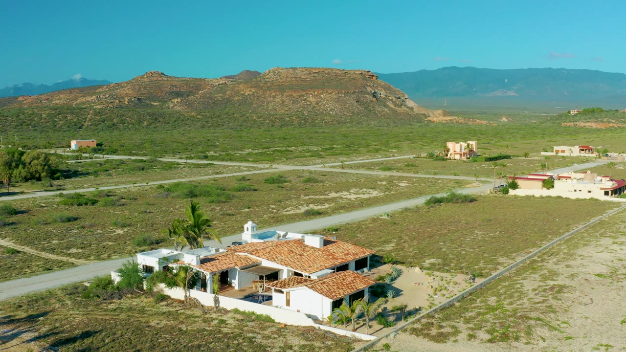 Wide aerial frame of homes, open green land, roads, hills, and blue sky.