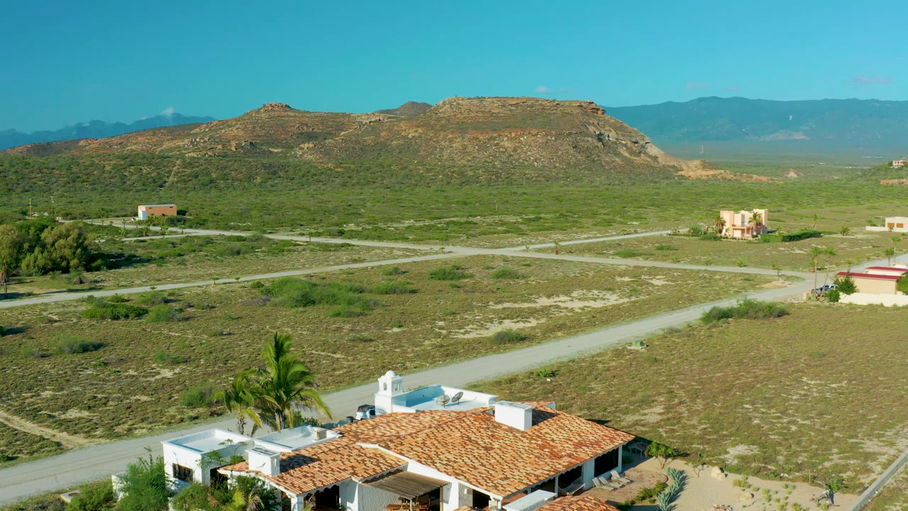 At 0:01, the La Ribera aerial view keeps the white house roof and palms at the bottom while pale dirt roads cross green-brown scrubland toward low hills, with no visible people.