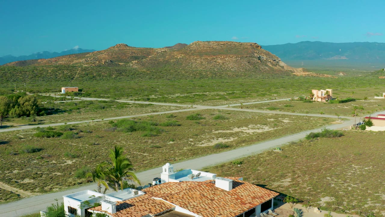 At 0:02, the frame shows more of the foreground roofline and palms below intersecting dirt roads, scattered homes, desert scrub, a flat-topped hill, distant mountains, and no visible people.