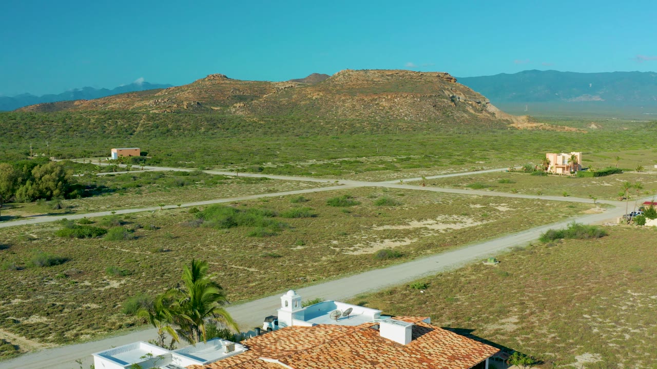 Open scrubland crossed by dirt roads with homes, palms, hills, mountains, and clear sky.