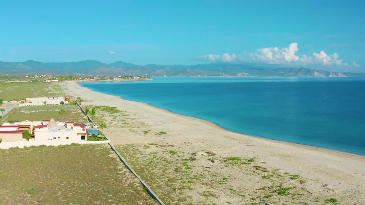 Opening aerial frame over La Ribera, Baja California Sur showing turquoise water, a pale sand beach, low scrubland, beachside homes, distant mountains, and no visible people.