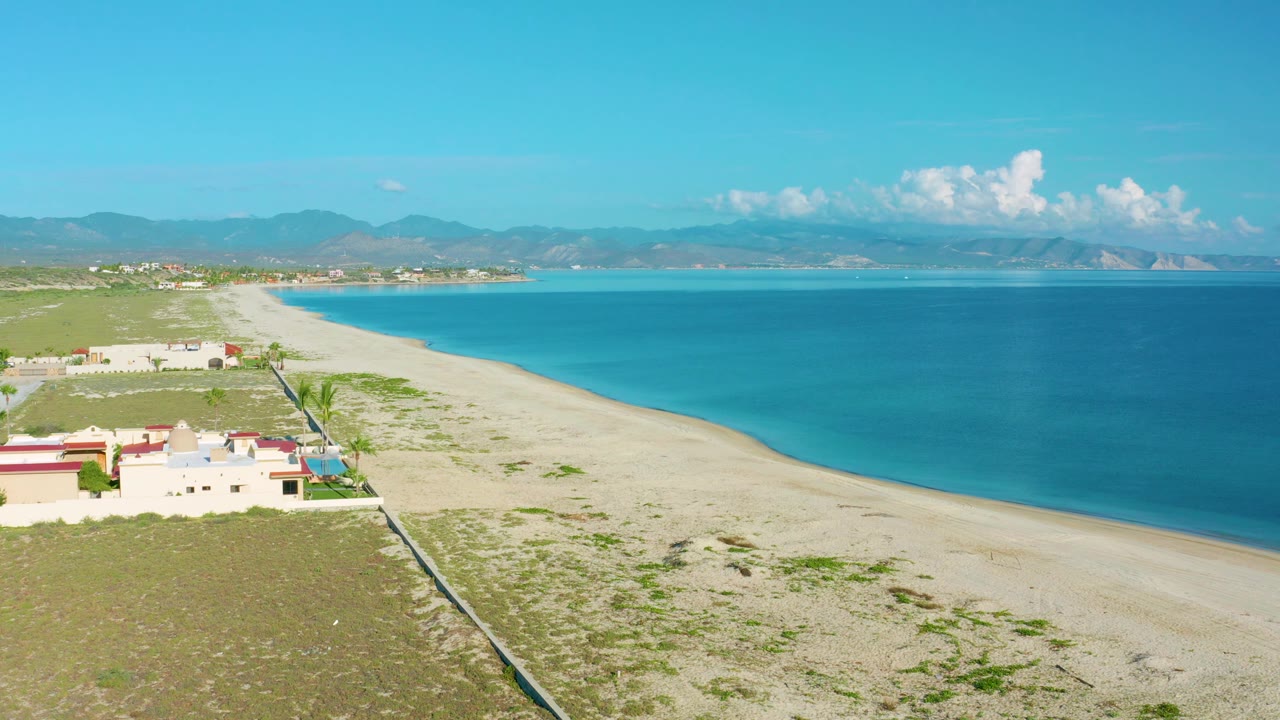 La Ribera aerial view with a curving pale beach, clear blue ocean, a cluster of low homes at the beach edge, mountain ridges in the background, and no visible people.