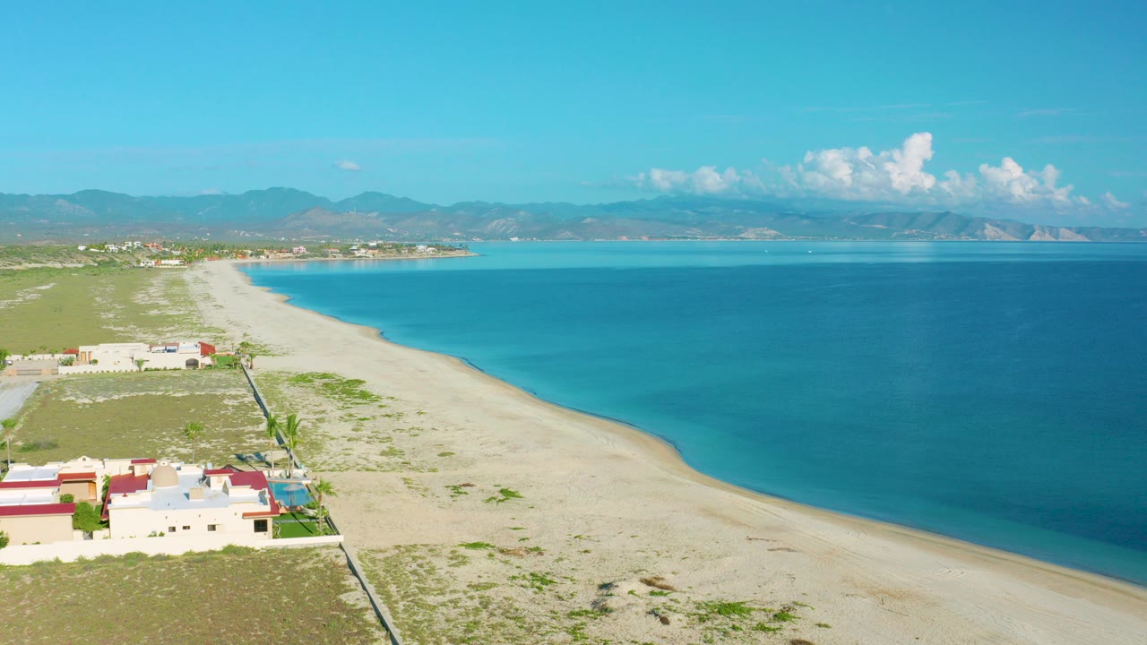 Opening aerial frame over La Ribera, Baja California Sur showing pale sand, blue water, beachside homes at lower left, distant mountains, white clouds, and no visible people.