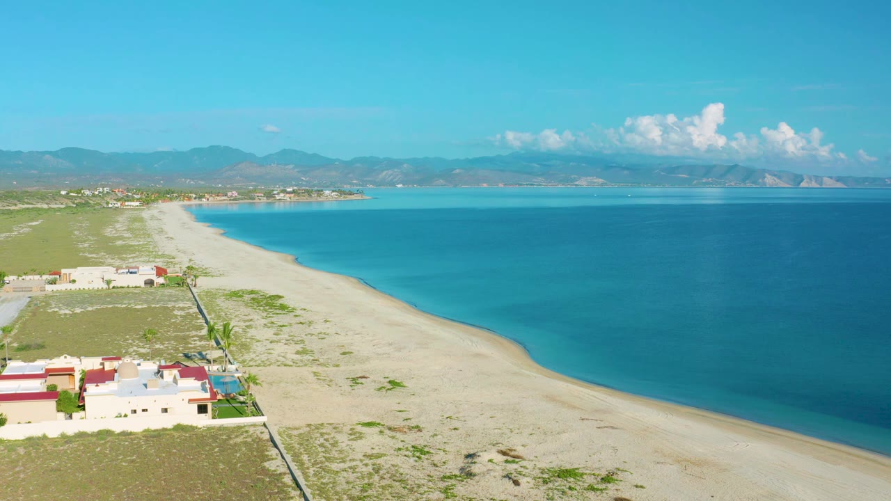 Aerial frame in La Ribera, Baja California Sur with the shoreline angled across the image, turquoise water on the right, beach homes at lower left, mountains on the horizon, and no visible people.