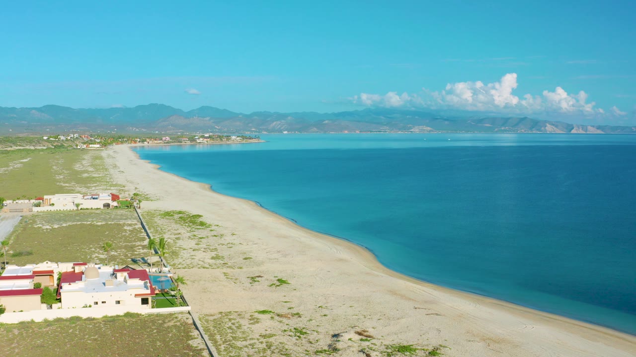La Ribera aerial frame showing a long pale beach beside deep blue water, a small cluster of beachside buildings in the foreground, mountain ridges across the horizon, and no visible people.