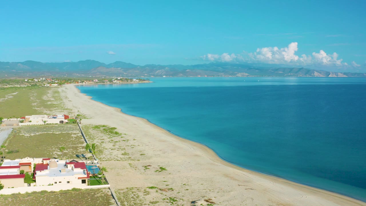 Aerial coastline with beach houses below, open sand, calm sea, and distant mountains.