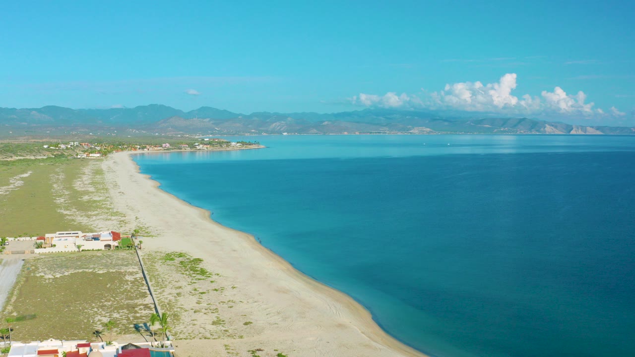 Aerial frame over La Ribera, Baja California Sur showing pale sand curving along bright blue water, low buildings at the lower left, distant mountains, clouds, and no visible people.