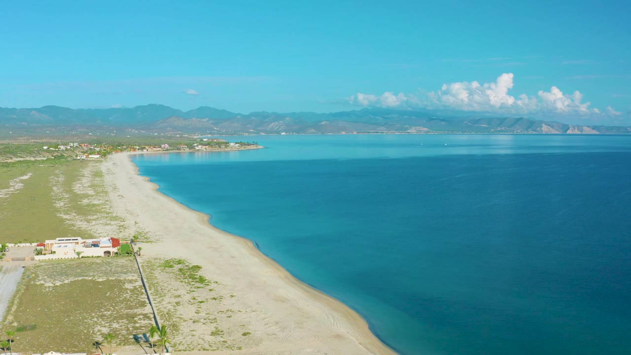 La Ribera aerial frame showing a diagonal strip of sand, deepening blue ocean to the right, small structures near the lower left edge, hazy mountains in back, and no visible people.