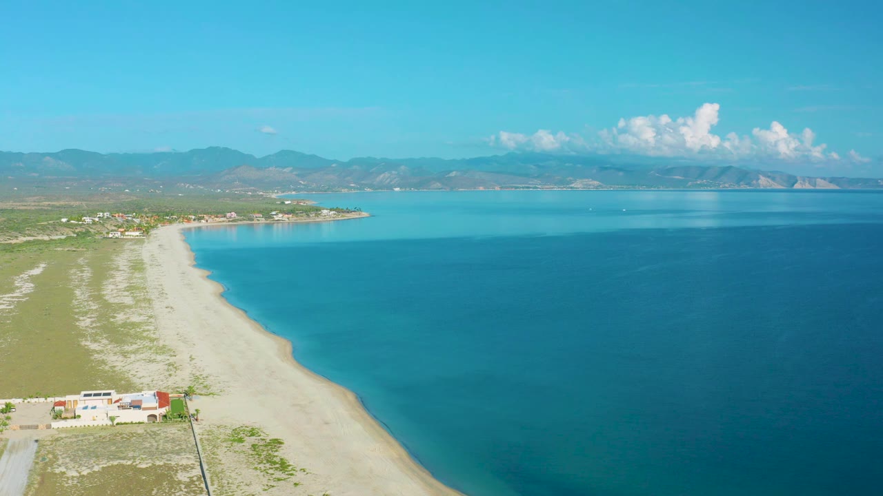 At 0:01, the La Ribera shoreline remains in a wide aerial view with low buildings near the lower edge, turquoise water to the right, mountains beyond the bay, and no visible people.