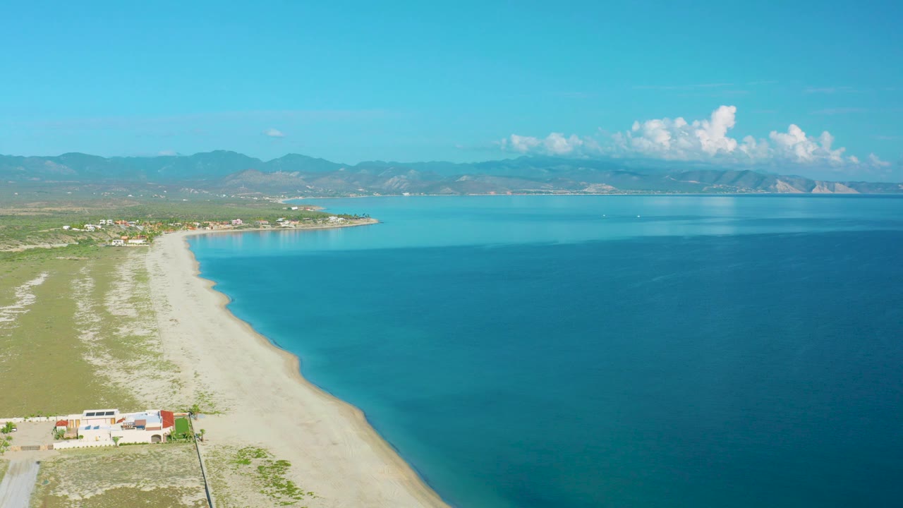 At 0:02, the beach curve and deep blue water dominate the La Ribera aerial frame, with green land on the left, mountain ridges in the distance, and no visible people.