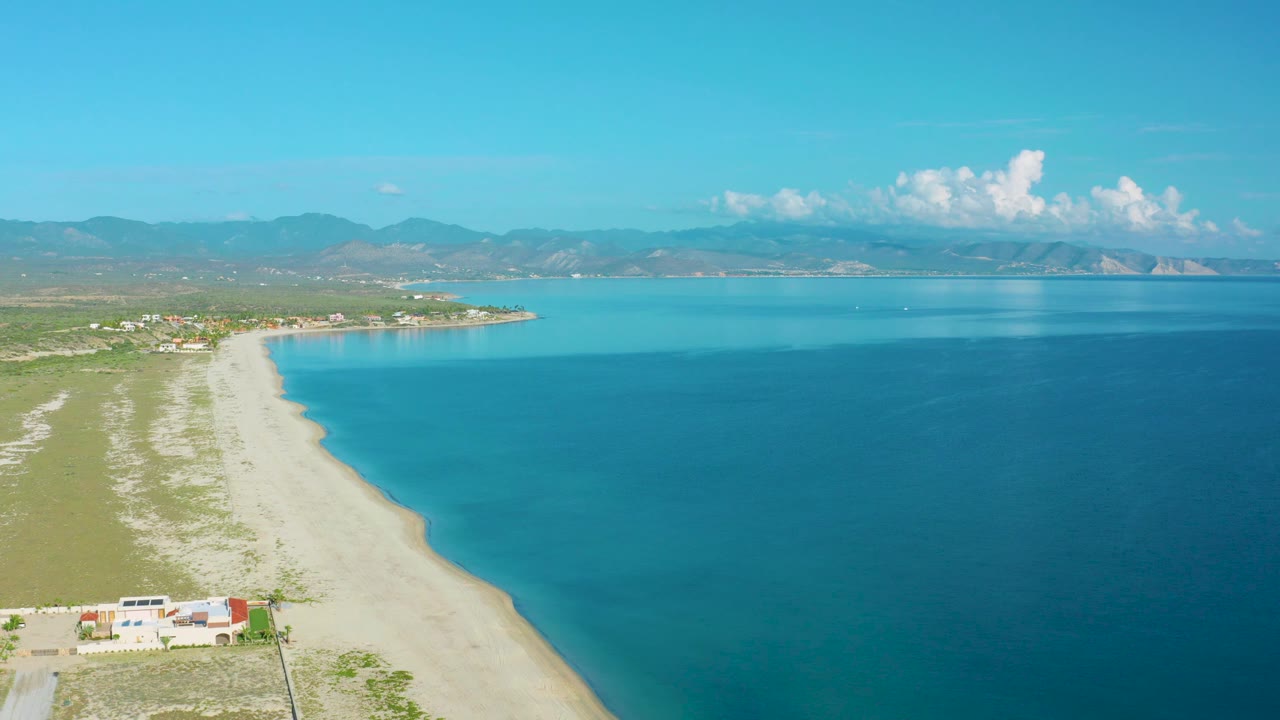 Elevated coastal view with beach, calm blue water, scrubland, and mountain horizon.