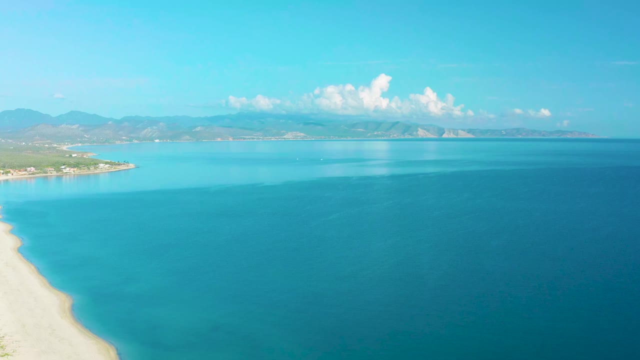 Opening frame over La Ribera, Baja California Sur with pale beach at lower left, wide blue sea, mountain ridge on the horizon, white clouds above, and no visible people.