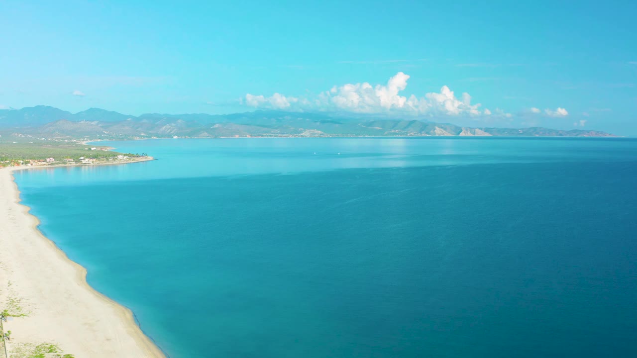 Aerial coastline with white sand at left and calm blue water stretching to mountains.