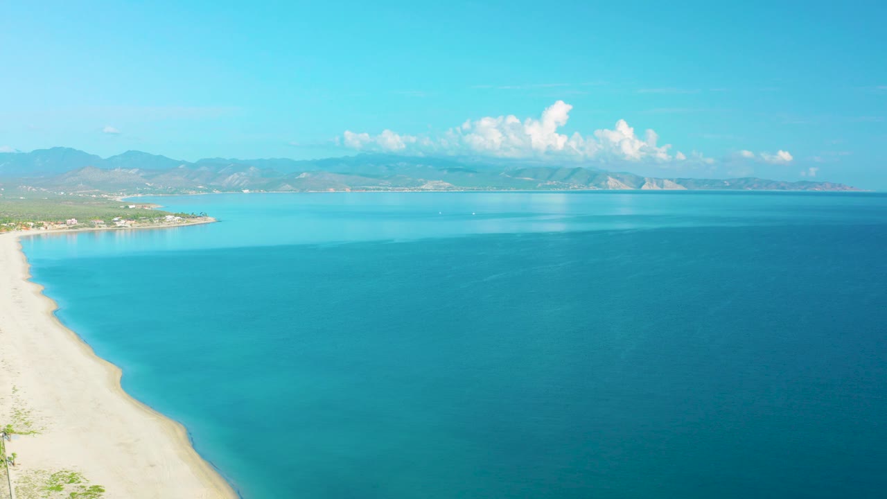 At 0:02, the La Ribera beach remains along the left side while turquoise water opens into a broad blue bay, with mountains and bright clouds beyond and no visible people.