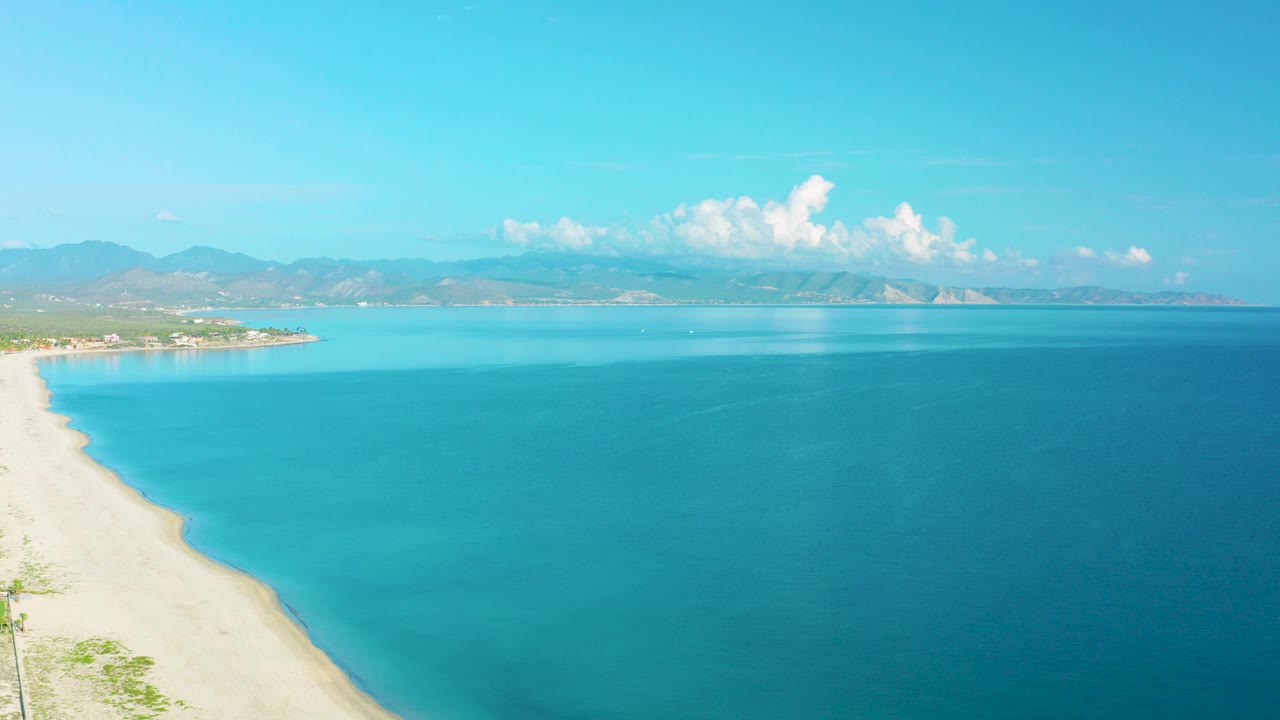Opening aerial frame over La Ribera, Baja California Sur, with turquoise water, a pale beach along the left edge, distant mountains, white clouds, and no visible people.