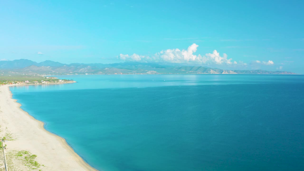 La Ribera aerial frame showing pale sand, shallow turquoise water near shore, darker open sea to the right, distant mountain ridges, and no visible people.