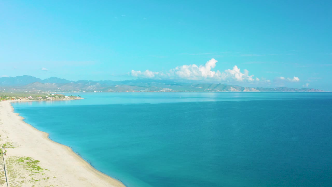 Opening aerial frame over La Ribera, Baja California Sur, with pale sand at left, turquoise water across the center, distant mountains on the horizon, white clouds, and no visible people.