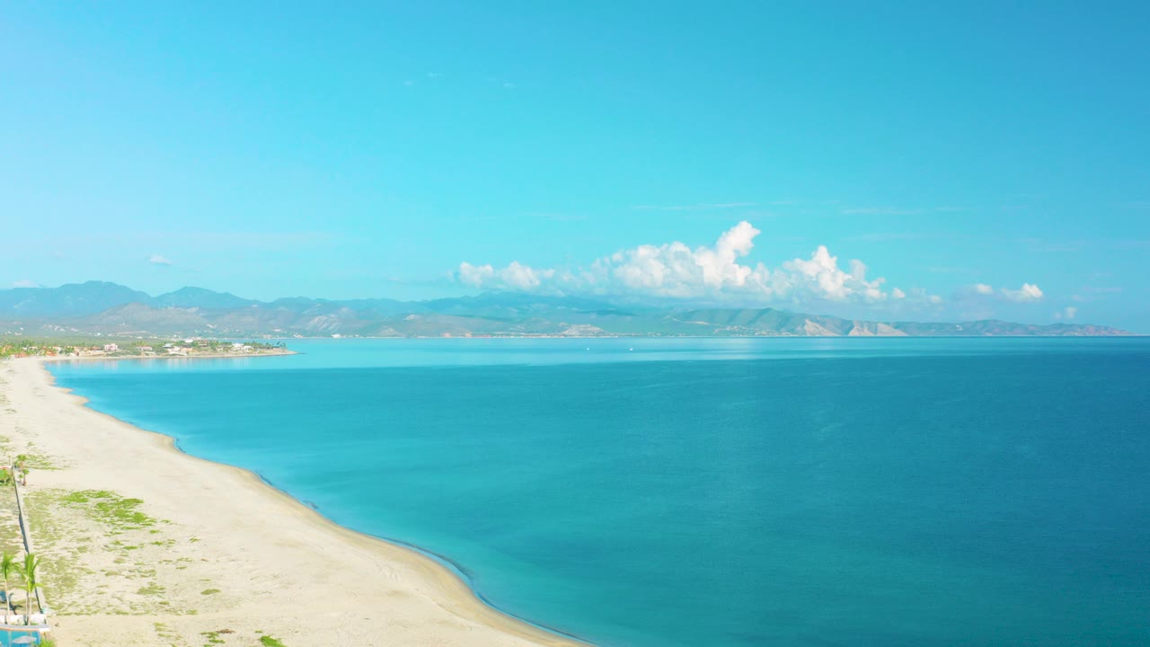 Opening aerial frame over La Ribera, Baja California Sur, with a pale sandy beach at left, turquoise water across the center, distant mountains on the horizon, and no visible people.