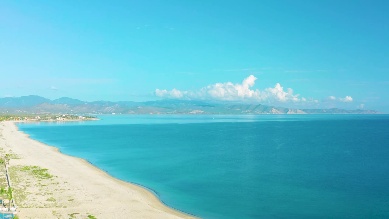 A later aerial view of La Ribera coastline shows the beach curve more clearly at left, broad turquoise water, low mountains in the distance, and no visible people.