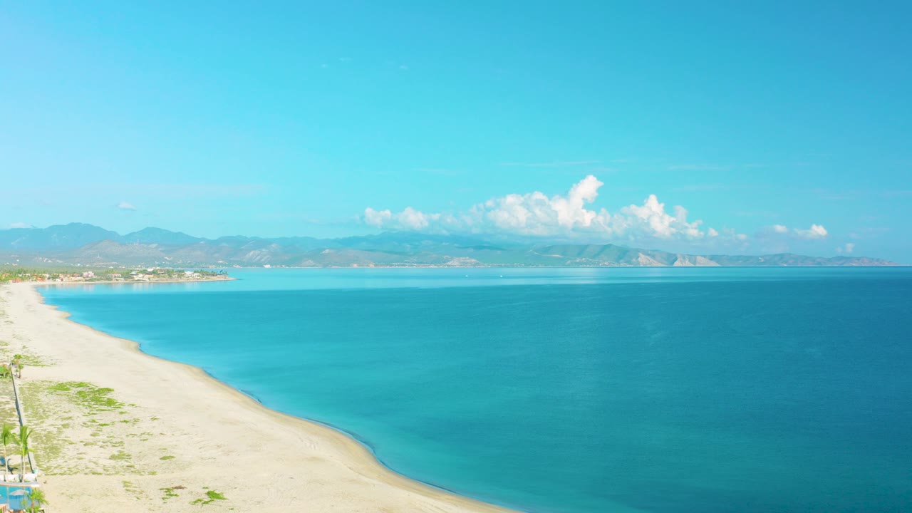 La Ribera beach remains along the left side while calm blue water fills the frame, backed by hazy mountains and a small cluster of white clouds, with no visible people.